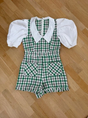 Grön och vit rutig kort byxdress - Superfin kort byxdress med grönt och vitt rutigt mönster, stora vita puffärmar och bred vit krage. Byxdressen har två fickor framtill och dragkedja i ryggen. Materialet känns som en mix av bomull och syntet, perfekt för dig som gillar statement-plagg.