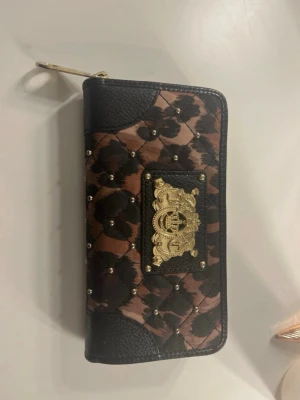 Juicy Couture plånbok med leopardmönster och gulddetaljer - Snygg och iögonfallande plånbok från Juicy Couture med quiltat leopardmönster, svarta detaljer och dekorativa guldfärgade nitar samt emblem på framsidan. En rymlig modell som passar bra för kort, sedlar och småsaker, och som ger en lyxig känsla till vardagen. Observera att delen man håller i för att öppna dragkedjan inuti saknas, men dragkedjan går fortfarande att öppna och stänga.