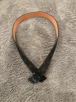 Svart  bälte - Säljer ett svart bälte  med klassiskt monogrammönster och silverfärgat spänne. Bältet är gjort i skinn med beige insida och har en exklusiv känsla. Perfekt accessoar för att lyfta vilken outfit som helst.