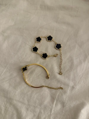 Svart och guld armbandsset - Säljer ett set med två armband i svart och guld. Ena armbandet har fem svarta fyrklöverformade hängen med guldkant, det andra är ett smalare guldarmband med ett svart fyrklöverhänge. Båda har justerbara kedjor för perfekt passform. Båda för 50 kr!!
