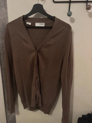 Brun kofta från Selected Homme - Snygg brun kofta från Selected Homme med v-ringning och knappar framtill. Långärmad modell i mjukt material, perfekt för lager på lager. Enkel och stilren design som passar till det mesta.