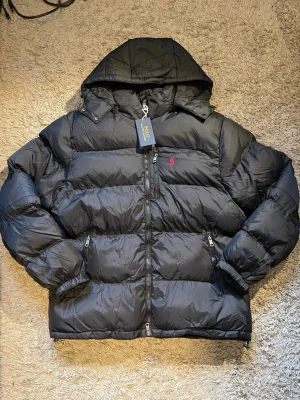 Svart pufferjacka från Polo Ralph Lauren - Svart pufferjacka från Polo Ralph Lauren med huva, dragkedja och två sidofickor med zip. Jackan har det klassiska röda Polo-logot broderat på bröstet och är tillverkad i polyester. Perfekt för kalla dagar och har en riktigt schysst oversized look.