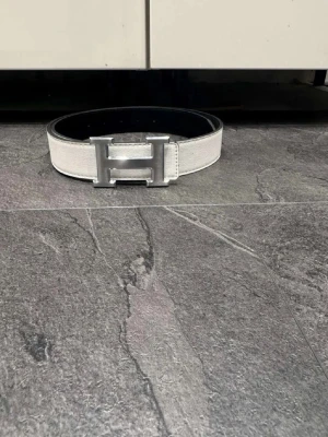 Vit och  skärp med H-spänne - Snyggt skärp i vitt och svart med ett stort metallfärgat H-spänne. Skärpet har en stilren design och är gjort i skinn med struktur. Perfekt accessoar för att lyfta din outfit.