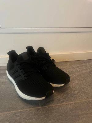 Svarta Adidas Ultraboost sneakers - Säljer ett par svarta Adidas Ultraboost sneakers med vit, responsiv sula och klassiska tre ränder på sidan. Ovandelen är i ventilerande mesh och skorna har snörning för perfekt passform. Perfekta för både träning och streetwear.