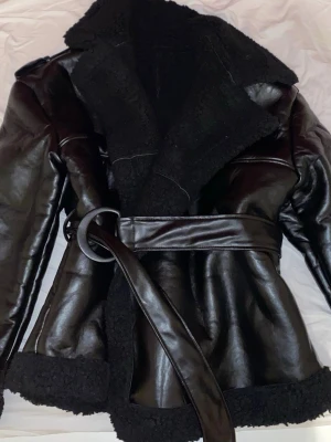 oversize Svart teddyjacka med bälte - Säljer en svart teddyjacka i skinnimitation med bred krage och mysigt teddyfoder. Jackan har ett matchande bälte i midjan och detaljer i samma material. Perfekt oversized look och riktigt varm för kyliga dagar.