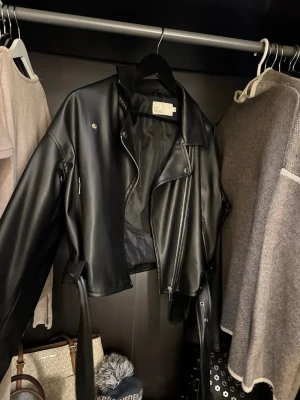Svart skinnjacka från NLY Trend - Säljer en svart skinnjacka från NLY Trend i storlek 32. Jackan är croppad med klassisk bikerstil, har dragkedja, knappar och bälte i midjan. Snygga detaljer som axelklaffar och fickor med dragkedja. Perfekt för dig som vill ha en edgy look.