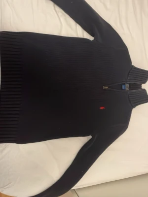 Svart stickad tröja Polo Ralph Lauren - Mörkblå stickad tröja från Polo Ralph Lauren med halv dragkedja och hög krage. Klassisk röd logga broderad på bröstet. Tröjan har ribbade muddar och nederkant, samt långärmad passform. Perfekt för kyliga dagar. 10/12 storlek