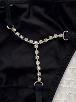 Svart stringtrosa med strassdetaljer - Svart stringtrosa med tunna band och glittrande strasskedja framtill som ger en extra lyxig känsla. Trosan har en enkel trekantsform och smala sidor, perfekt för dig som vill sticka ut lite extra.