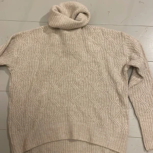 Beige stickad polotröja - Mysig beige stickad polotröja med ett snyggt kabelstickat mönster. Perfekt för kyliga dagar med sin höga krage och långa ärmar. Tröjan har ribbade muddar vid ärmslut och nederkant för extra komfort.