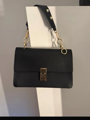 Svart axelväska med gulddetaljer - Stilren svart axelväska i skinn med bred axelrem och snygga guldfärgade metalldetaljer. Väskan har ett klassiskt, fyrkantigt format och ett elegant spänne framtill. 🤍