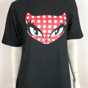 Svart t-shirt med kattmotiv och prickar L - Cool svart t-shirt med ett stort kattansikte på bröstet. Katten har röda och vita prickar och markerade ögon, vilket ger en unik och lekfull stil. Perfekt för dig som vill sticka ut med din outfit. Stl L. Helt ny.