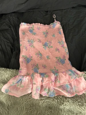 Rosa blommig volangkjol - Supersöt kort kjol i ljusrosa med blommigt mönster i blått, grönt och lila. Kjolen har smockad midja och bred volang längst ner som ger en luftig och romantisk känsla. Perfekt för varma dagar och somriga outfits. Säljer nu under vintersäsongen och därför är den billig. 💓