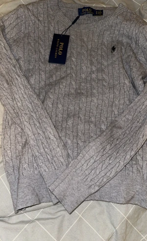 Grå kabelstickad tröja Polo Ralph Lauren - Barn storlek S Säljer en grå kabelstickad tröja från Polo Ralph Lauren med rund halsringning och diskret logga på bröstet. Tröjan har långa ärmar och ribbade muddar vid ärmslut och nederkant. Perfekt för lager på lager under hösten och vintern.