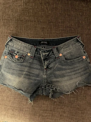 True Religion Joey Cut-Off jeansshorts - Säljer ett par grymma blå jeansshorts från True Religion, modell Joey Cut-Off. De är lågmidjade med råa, fransiga kanter och har röda detaljer på bakfickorna. Klassisk femficksdesign och coola kopparfärgade knappar. Perfekta för sommaren! Har bara använt ett fåtal gånger 