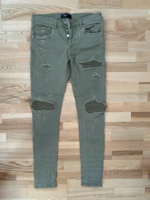 Olivgröna skinny jeans från Amiri - Säljer ett par olivgröna skinny jeans från Amiri med slitna detaljer och ribbade lappar vid knäna. Byxorna har knappgylf, fem fickor och klassisk jeanslook. Perfekta för dig som gillar streetwear och vill ha en edgy stil.