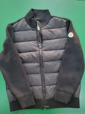 Svart pufferjacka från Moncler - Snygg svart pufferjacka från Moncler med stickade ärmar och quiltad front. Jackan har hög krage, dragkedja framtill och Moncler-logga på ena ärmen. Perfekt för dig som vill ha en stilren och sportig look.