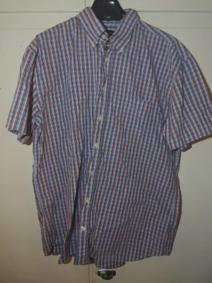 Rutig kortärmad skjorta från GANT XL - Säljer en klassisk rutig skjorta från GANT i storlek XL. Skjortan har korta ärmar, knappar framtill och en bröstficka. Färgerna är blå, röd och vit, och materialet är bomull. Perfekt för dig som gillar en avslappnad men ändå stilren look.