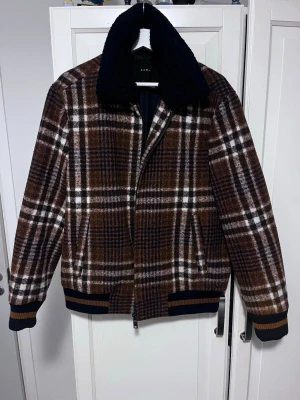 Jacka - Säljer en riktigt snygg teddyjacka från Zara med rutigt mönster i brunt, vitt och mörkblått. Jackan har en stor svart teddykrage, dragkedja framtill och ribbade muddar med bruna detaljer. Perfekt för dig som vill ha en varm och trendig jacka med retrovibbar.