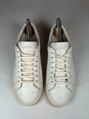 Vita sneakers från Saint Laurent - Clean och stilrena vita sneakers från Saint Laurent med diskret logga i guld på sidan. Skorna har klassisk låg profil, rund tå och platt sula. Tillverkade i mjukt skinn med vita snören och minimalistisk design som passar till allt.