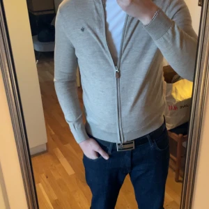 Beige kofta med dragkedja från Morris - Säljer en beige kofta från Morris med dragkedja framtill och diskret logga på bröstet. Tröjan är långärmad, har ribbade muddar och är tillverkad i mjuk bomull. Perfekt för lager på lager under höst och vår. 