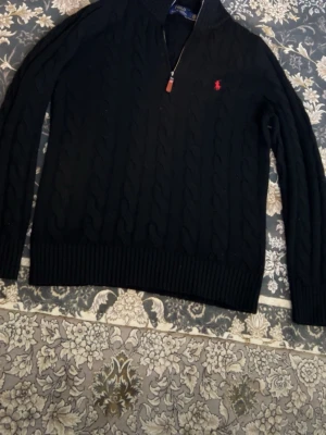 Svart kabelstickad tröja Polo Ralph Lauren - Kabelstickad svart tröja från Polo Ralph Lauren med halv dragkedja och röd broderad logga på bröstet. Tröjan har ribbade muddar, hög krage och är långärmad. Perfekt för kyliga dagar och snygg till jeans eller chinos.