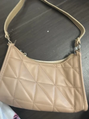 Beige quiltad axelväska - Säljer en beige axelväska i quiltad design med triangulära sömmar. Väskan har ett kort axelband och stängs med dragkedja. Perfekt storlek för det viktigaste och ger en clean look till din outfit.