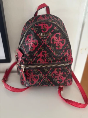 Svart och röd ryggsäck från Guess - Cool miniryggsäck från Guess med svart botten och röda samt guldiga logomönster över hela väskan. Väskan har röda axelband, dragkedja i silver och flera fack både inuti och utanpå. Perfekt för dig som vill ha en snygg och praktisk accessoar.