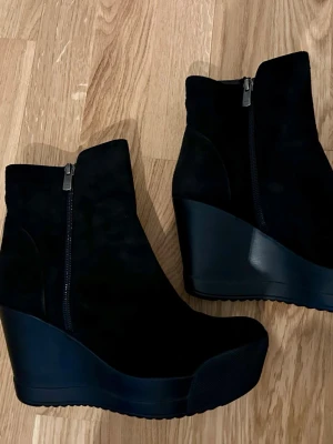 Svarta kilklacksstövletter i mocka - Säljer ett par svarta stövletter med kilklack(plattform) och dragkedja på sidan. Skorna har en rund tå och är tillverkade i mjuk mocka med en hög platåklack. Perfekta för dig som vill ha både höjd och komfort i din look.