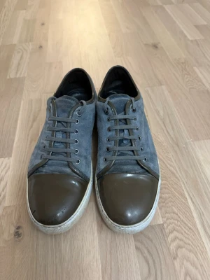 Lanvin - Säljer ett par stilrena sneakers från Lanvin i grå mocka med mörkbruna detaljer och tåparti i blankt skinn. Skorna har rund tå, snörning och vit sula. Perfekta för dig som gillar en clean och exklusiv look. Passar till det mesta.