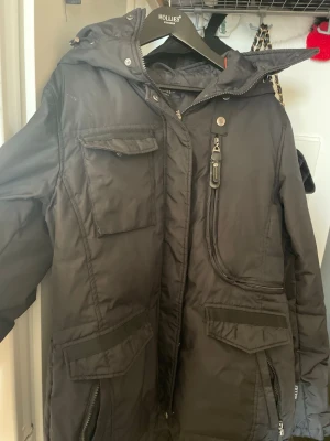 Svart parkas från Hollies - Säljer en svart parkas från Hollies med huva och flera praktiska fickor framtill. Jackan har dragkedja, knappar och detaljer som ger en cool look. Perfekt för kalla dagar och har en längre modell som skyddar mot vind.