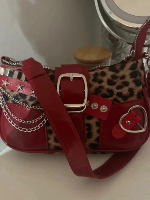 Röd axelväska med leopardmönster - Unik röd axelväska med leopardmönstrade detaljer och coola silverkedjor. Väskan har stora spännen, nitar, stjärnor och ett hjärta i metall. Materialet är glansigt syntetläder och dragkedja upptill. Perfekt för dig som vill sticka ut med din stil.
