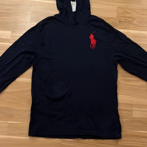 Ralph Lauren - Marinblå hoodie från Ralph Lauren med stor röd logga på bröstet. Tröjan har huva och långa ärmar, perfekt för en chill och stilren look. Tillverkad i mjuk bomull som känns skön mot huden. Storlek S. Den är använd men inga defekter. Ser som ny ut men använd. Kan gå ner lite i pris vid annan affär