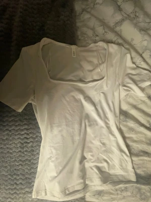 Vit topp med fyrkantig ringning H&M - Vit topp från H&M i storlek S med korta ärmar och snygg fyrkantig ringning. Tillverkad i mjuk bomull, perfekt för en clean och enkel look. Passformen är normal och toppen är helt enfärgad utan mönster eller detaljer.
