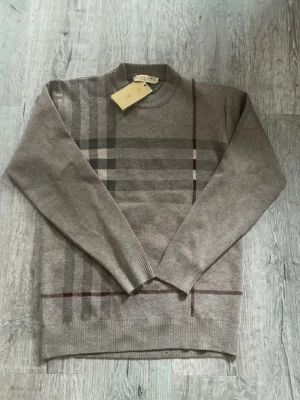 Beige rutig stickad tröja Burberry - Snygg beige stickad tröja från Burberry med klassiskt rutmönster i grått och vinrött på framsidan. Rund hals och långa ärmar med ribbade muddar. Perfekt för dig som gillar stilrena och tidlösa plagg med lyxig känsla.