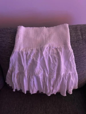 Vit volangkjol med smock - Supersöt vit kortkjol med smock i midjan och volangdetaljer nertill. Kjolen är gjord i ett lätt bomullstyg som känns luftigt och passar perfekt till varma dagar. Enkel att matcha med både toppar och sneakers för en chill look.