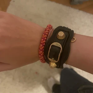 Balenciaga svart läderarmband - Säljer ett svart armband från Balenciaga i äkta läder med snygga guldfärgade detaljer och spänne. Armbandet har en cool och chunky design som sticker ut och passar perfekt för dig som gillar statement-accessoarer.