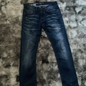 Mörkblå jeans från Smog Denim - Snygga mörkblå jeans från Smog Denim med slitna detaljer och kontrastsömmar. Klassisk femficksmodell med coola ljusa tvättade partier framtill. Perfekta för dig som gillar en avslappnad men trendig look. Storlek 30/32