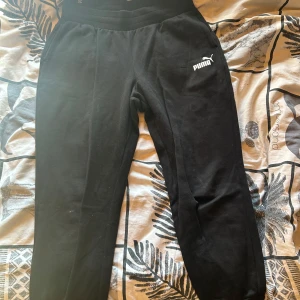 Svarta mjukisbyxor från Puma - Svarta mjukisbyxor från Puma med vit logga på vänster lår. Byxorna har elastisk midja och är tillverkade i mjukt material som passar perfekt för chill eller träning. Klassisk och enkel design med smalare ben nertill. Lite kortare i längden.