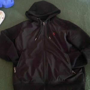 Svart hoodie från Polo Ralph Lauren M - Svart hoodie från Polo Ralph Lauren med dragkedja framtill och huva med snörning. Klassisk röd logga broderad på bröstet och två fickor med dragkedja. Perfekt för chill dagar eller när du vill ha en clean streetstyle.