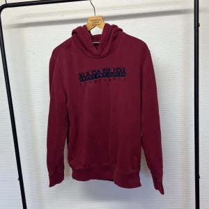 Vinröd hoodie från Napapijri - Säljer en vinröd hoodie från Napapijri med broderad logga på bröstet. Tröjan har huva och är tillverkad i mjuk bomull, perfekt för chill dagar. Klassisk passform och långa ärmar. Snygg och enkel att matcha med jeans eller joggers.