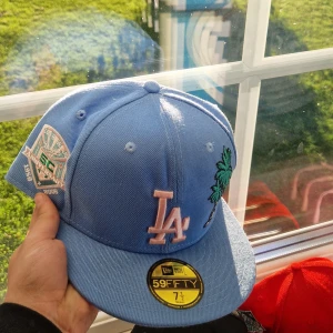 Blå LA New Era 59FIFTY keps - Snygg ljusblå 59FIFTY keps från New Era med LA-logga i rosa framtill och broderad palm på sidan. Kepsen har även en patch på sidan och klassisk flat skärm. Perfekt för dig som gillar streetwear och sportig stil.