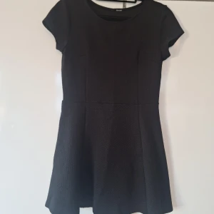 Svart kortärmad klänning från Ginatricot - En stilren svart klänning från Ginatricot med korta ärmar och rund halsringning. Klänningen har en lätt utställd kjol och är sydd i ett diskret strukturerat tyg. Perfekt för dig som gillar enkel och klassisk stil.