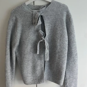 Grå ribbad kofta med knyt - Mysig grå ribbstickad kofta med långa ärmar och två knytband framtill. Perfekt för lager-på-lager och enkel att styla till olika outfits. Koftan har en klassisk rund halsringning och är tillverkad i ett mjukt stickat material.