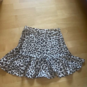 Leopardmönstrad volangkjol från New Yorker - Snygg kjol från New Yorker med leopardmönster i svart och beige. Kjolen har volang nertill som ger en extra touch och är i ett lätt, glansigt material. Finns dragkedja på sidan😊