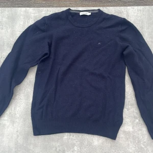 Mörkblå ulltröja från J.Lindeberg - Liten i storlek därför lade jag strl XS.                    Snygg mörkblå stickad tröja från J.Lindeberg med diskret logga på bröstet. Klassisk rund halsringning, ribbade muddar och nederkant. Perfekt för lager-på-lager och passar till allt. Materialet är mjuk ull som håller dig varm under kyliga dagar.