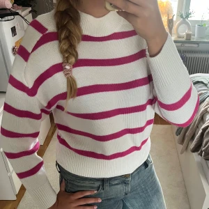 Vit och rosa randig stickad tröja - Säljer en vit stickad tröja med rosa ränder. Tröjan har rund halsringning, ribbade muddar och är långärmad. Perfekt för dig som gillar färg och vill ha en mysig men ändå snygg look. Passar till jeans eller kjol.