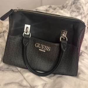 Svart handväska från Guess med logomönster - Snygg svart handväska från Guess med klassiskt logomönster och silverfärgade detaljer. Väskan har två handtag, dragkedja upptill och rymligt fack inuti. Tillverkad i syntetmaterial med stilren design som passar perfekt till din outfit.
