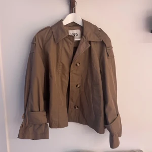 Beige trenchcoat från Zara -  beige trenchcoat från Zara med klassisk krage, knappar framtill och  detaljer vid axlar och ärmslut. Vill man att ärmslutet ska vara mer utsvängt kan man använda knapp längre ut än vad som används på bild 3. 