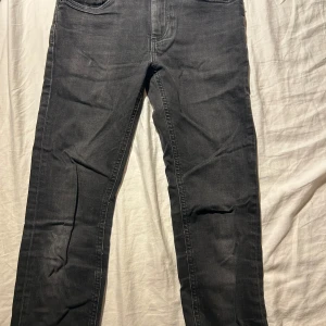 Svarta jeans från Tommy Hilfiger - Svarta jeans från Tommy Hilfiger med klassisk femficksdesign och diskret logga på bakfickan. Jeansen har raka ben och är tillverkade i mjukt bomullstyg med en snygg mörk tvätt. Perfekta för en stilren och avslappnad look.