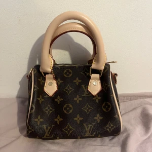  handväska - Snygg handväska. Väskan är brun med beiga handtag och detaljer, har guldfärgad dragkedja. Perfekt storlek för det viktigaste och en riktig statement piece.